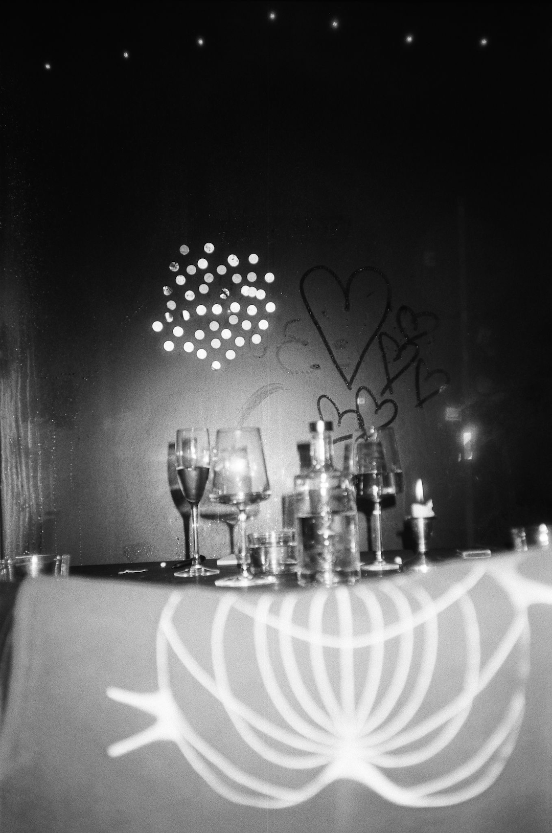 A black and white photograph capturing a dimly lit, intimate table setting. The table is adorned with several glasses, bottles, and a lit candle, creating a cozy ambiance. In the background, a foggy window displays drawings of hearts and abstract shapes, possibly made by someone using their finger. Small dots of light are visible through the window, adding to the warm and inviting atmosphere. The foreground features a cloth with a simple, elegant pattern. The overall scene evokes a sense of romance and quiet celebration.