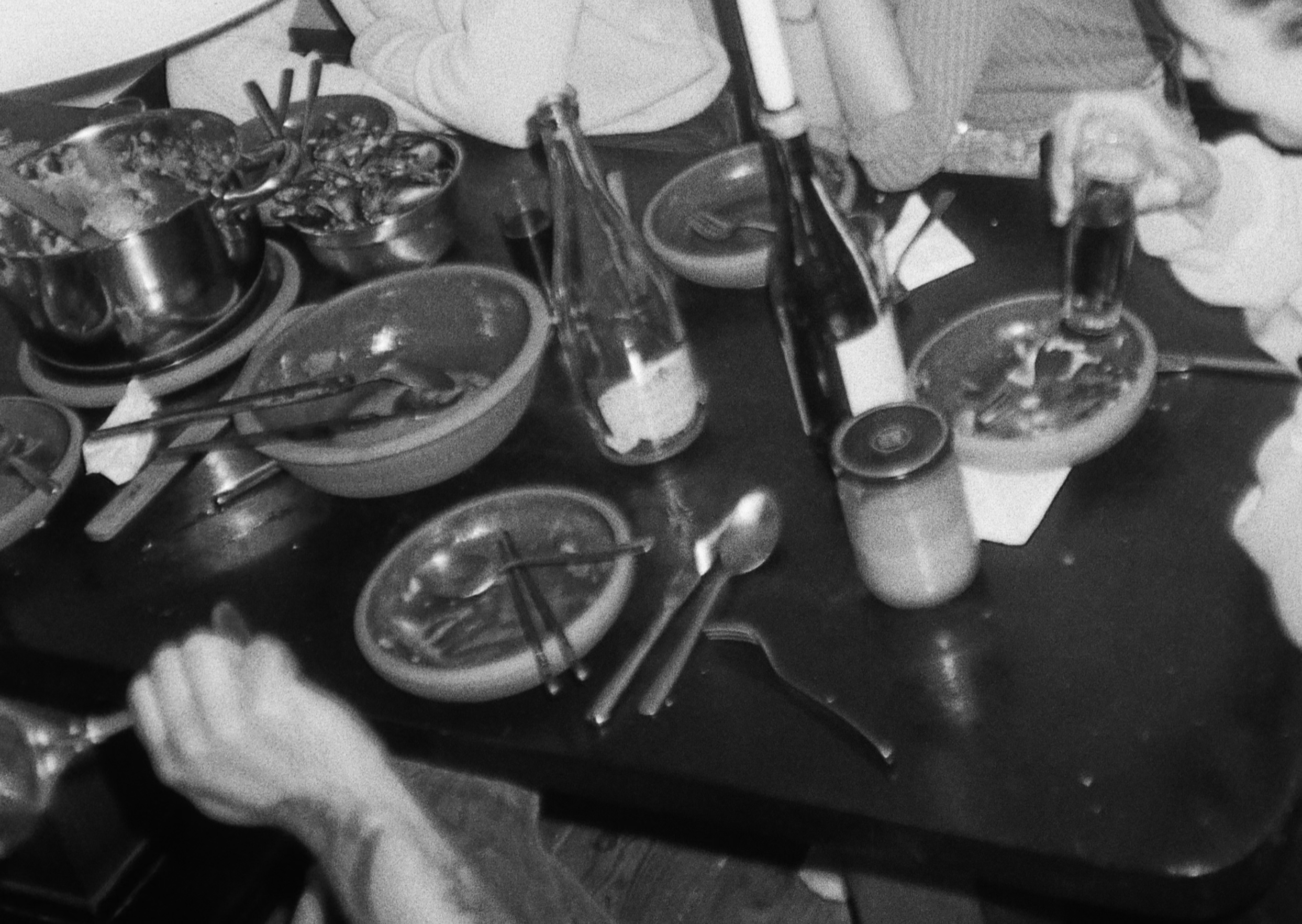 A black and white photograph of a dining table after a meal. The table is cluttered with bowls, pots, utensils, bottles, and partially eaten food. Various dishes and drinks are scattered across the table, indicating a recently concluded meal. Several people are partially visible, engaged in conversation or finishing their drinks. The atmosphere is informal and lively, with a focus on the remnants of the meal and the social interaction around the table.
