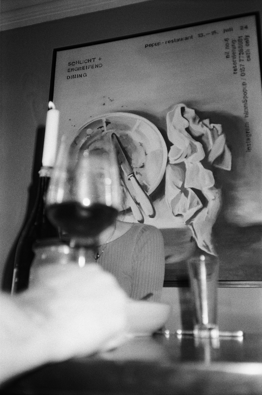 A black and white photograph taken in a dining setting. The focal point is a person sitting at a table, obscured by a wine glass and candle in the foreground. The background features a poster with the text 'SCHLICHT + ERGREIFEND DINING' and other information about a pop-up restaurant event. The poster also displays a plate, utensils, and a crumpled napkin. The image has a casual, candid feel with a blurred depth of field.